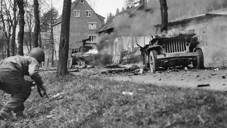 一名美国士兵小心翼翼地靠近被德军伏击摧毁的两辆燃烧的汽车。作为一名侦察兵，为了在战场上保持机动性，二等兵塞维尔从不穿装备或厚衣服。